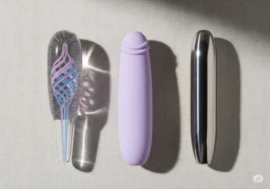 A flat lay of three affordable body-safe dildos on a linen background, showcasing different materials: a glass dildo with a pink and blue swirl, a matte purple silicone dildo, and a polished stainless steel dildo.
