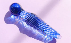 Artistic shot of a body-safe glass dildo for beginners in blue with intricate patterns, glowing under soft light reflections on a pink surface.