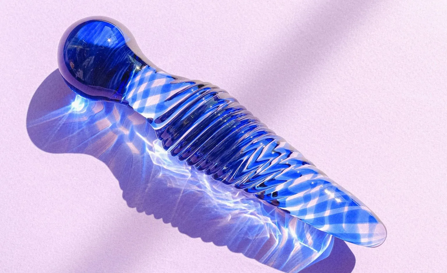 Artistic shot of a body-safe glass dildo for beginners in blue with intricate patterns, glowing under soft light reflections on a pink surface.