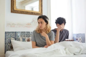 A couple sits together in a bedroom, experiencing a moment of misunderstanding while learning how to improve intimacy after erectile dysfunction.