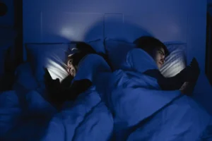 A couple lying in bed using smartphones at night, illuminated by screen light, showing the impact of sleep quality on sexual desire.