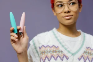 A young woman with eyeglasses holds two colorful discreet small vibrators for women against a purple background.