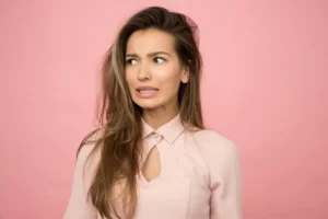 A young woman with a playful, uncertain expression illustrating the role of pheromones in female arousal myths vs facts against a pink background.
